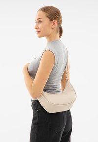 Borsa hobo in pelle beige con una texture liscia, forma curva e dettaglio del logo dorato. Indossata a tracolla sopra un top grigio e jeans neri.