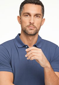 Camisa polo azul marino con un acabado texturizado, que presenta un collar de dos botones y mangas cortas, exhibida en un modelo con una expresión neutral.