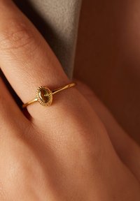 Gouden ring met een ovale groene steen, een gestructureerde band en ingewikkelde pareldetails rondom de steen, gedragen op een hand.