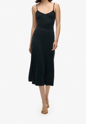 Zwarte midi-jurk met dunne spaghettibanden, aansluitende bustier en uitlopende zoom. Gemaakt van een gladde stof met een zachte textuur, gedragen met tan sandalen.