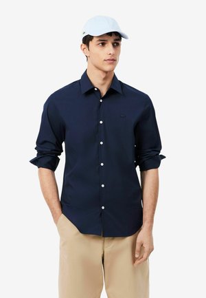 Giovane uomo con indosso una camicia blu navy a maniche lunghe abbottonata, pantaloni beige e un cappellino da baseball bianco, con le mani in tasca.