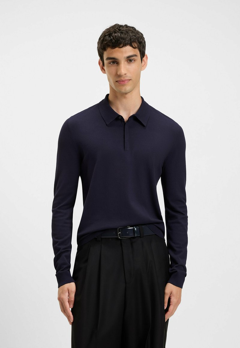 Młody mężczyzna w granatowej, długorękawowej koszulce polo, włożonej w czarne spodnie w kant, z czarnym paskiem, stojący na jednolitym białym tle.