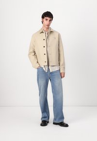 Beige lerretjakke med svarte knapper, båret over en lys rutete skjorte, sammen med lyseblå denimjeans og svarte sko. Enkelt design.