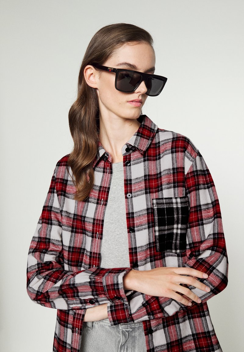 Czerwona i szara koszula flanelowa w kratę z czarną kieszenią, noszona na szarej koszulce, w połączeniu z dużymi czarnymi okularami przeciwsłonecznymi. Gładkie włosy w luźnych falach.