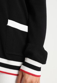 Svart tröja med vita ribbade manschetter och ficka. Manschetterna har röda, vita och svarta ränder. Mjuk textur, avslappnad design.