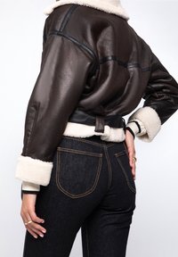 Brun läderjacka i kort modell med fårskinnskrage och manschetter, svart piping och detaljer med bälte vid midjan, tillsammans med mörka denimjeans.