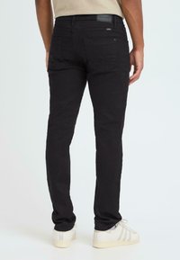 Crne traperice uskog kroja s stražnjim džepovima i teksturiranom tkaninom. Uparene s bež majicom i bijelim tenisicama.