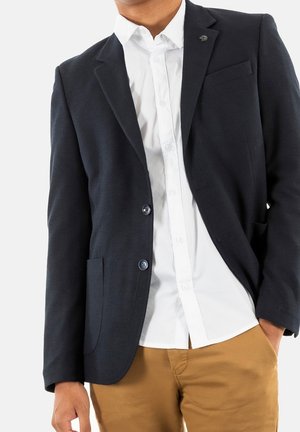 Mężczyzna w czarnej marynarce na białej koszuli i beżowych spodniach, stojący z jedną ręką w kieszeni na białym tle.