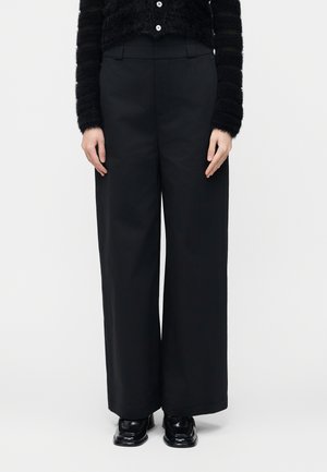 Nő magas derekú, bő szárú fekete nadrágot visel fekete, fényes, négyzet alakú orrú cipőkkel és fekete, bolyhos, gombos pulóverrel.