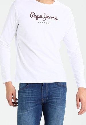 Homme portant une chemise blanche à manches longues avec le texte « Pepe Jeans London » et un jean en denim bleu, debout devant un fond uni.