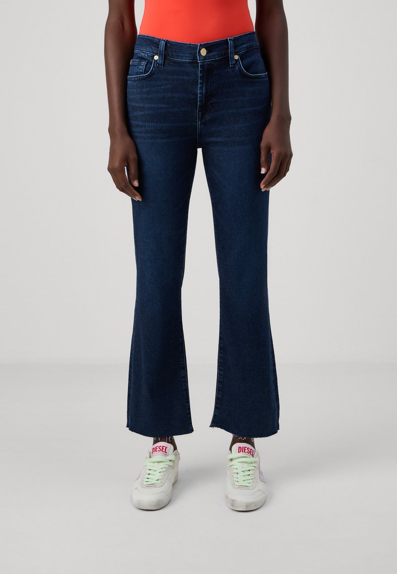 Donkerblauwe flared jeans met een hoge taille, denimtextuur en subtiele naaddetails. Draag met witte sneakers met roze accenten.
