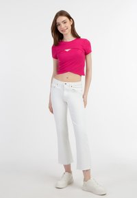 Feszes, rózsaszín crop top elülső kivágással, fehér harangnadrággal párosítva. A modellen fehér sportcipő van. Egyszínű fehér háttér.
