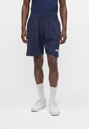 Männliches Model trägt marineblaue Freizeithosen mit weißem Logo, kombiniert mit weißen Sportsocken und weißen Turnschuhen vor weißem Hintergrund.