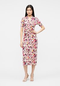 Femeie în picioare, cu mâna pe șold, purtând o rochie midi mulată, cu imprimeu abstract multicolor în roz, negru și galben, și pantofi bej cu toc.