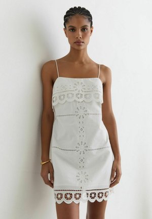 Donna con i capelli intrecciati che indossa un vestito bianco senza maniche con motivi floreali traforati, in piedi davanti a una parete bianca liscia.