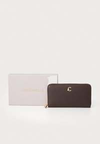 Portofel din piele maro cu un finisaj texturat, hardware auriu și un logo. Prezentat alături de o cutie roz deschisă etichetată "Coccinelle."