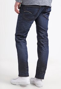 Vaqueros de mezclilla azul oscuro con un corte slim, que presentan un diseño clásico de cinco bolsillos y sutiles acentos de costura. Combinados con zapatillas blancas.