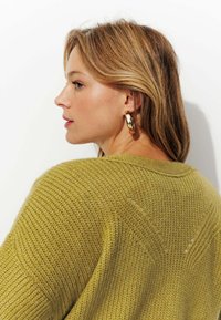 Pull en tricot de couleur vert olive avec un motif texturé, présentant un décolleté en V et une légère épaule tombante. Boucles d'oreilles en or et cheveux lisses.