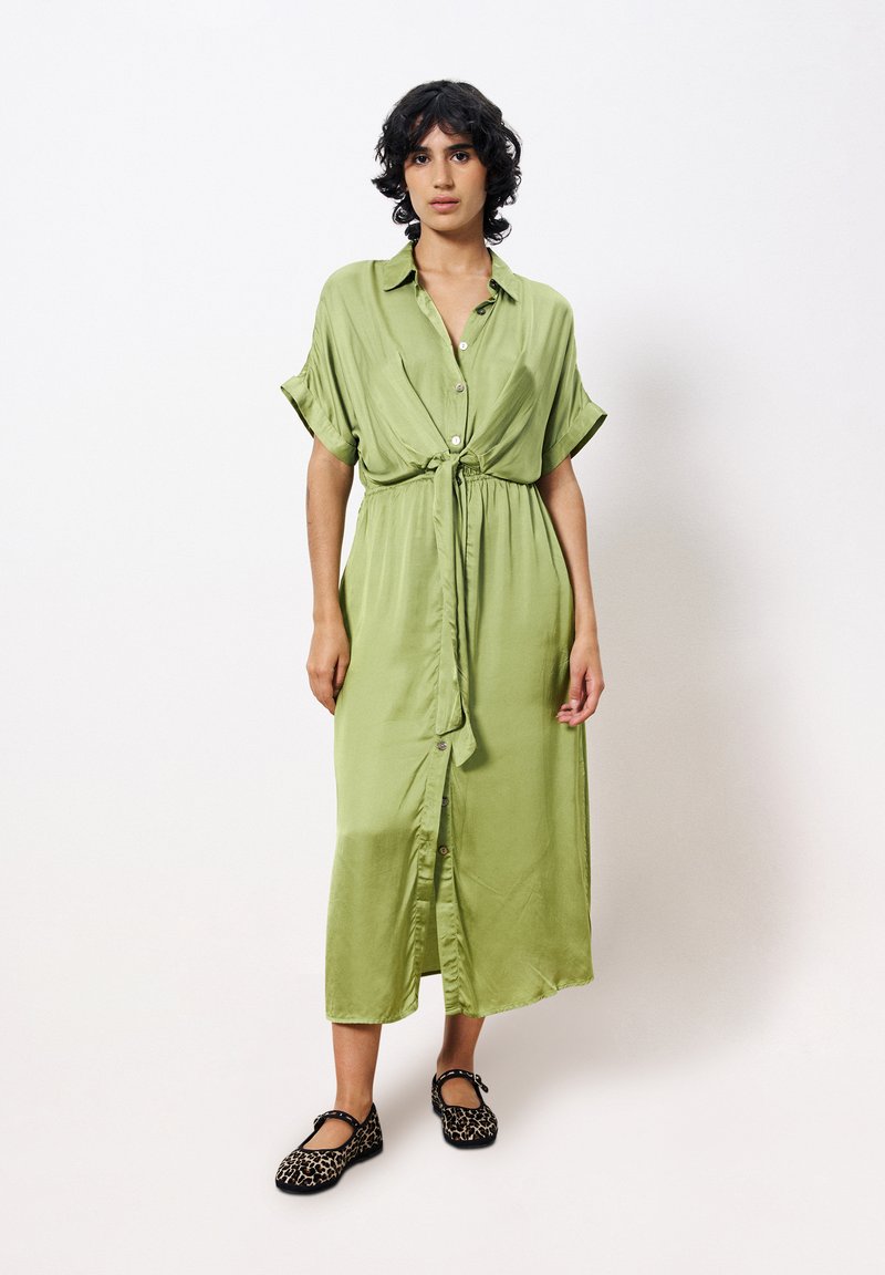 Grünes Satin-Midikleid mit kurzen Ärmeln, einer Knopfleiste und einem Taillenband. Kombiniert mit schwarzen Ballerinas im Leopardenprint.