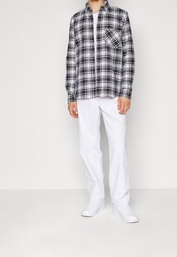 Camicia a scacchi bianca e nera con una singola tasca sul petto, abbinata a pantaloni bianchi e sneakers bianche. Linee pulite, stile casual.