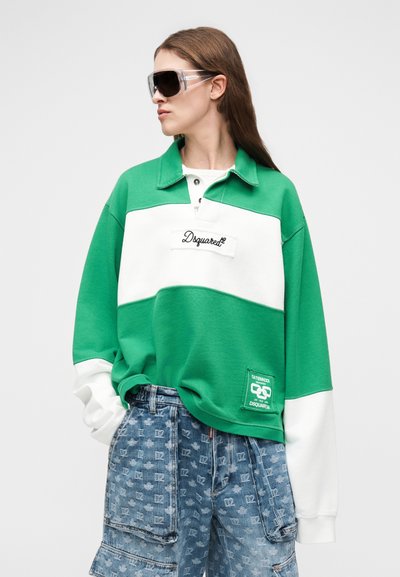 Žena odjevena u zeleni i bijeli Dsquared2 pulover s logotipom i uzorkirane traperice cargo kroja, gleda u stranu s velikim sunčanim naočalama.