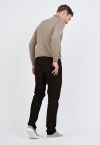 Vaalea ruskea neulepusero, mustat corduroyhousut ja harmaat lenkkarit. Näkymä takaa, joka näyttää materiaalien istuvuutta ja tekstuuria.