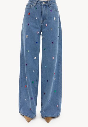 Blauwe jeans met wijde pijpen, versierd met meerkleurige, edelsteenachtige studs, gedragen met beige schoenen met puntige neus.