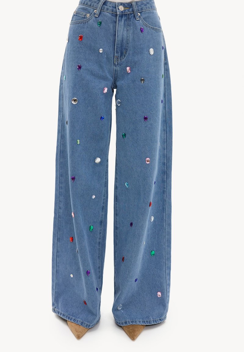 Vaqueros azules de pierna ancha adornados con tachuelas multicolores que parecen piedras preciosas, combinados con zapatos beige de punta afilada.