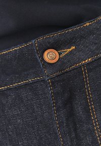 Sötét kék farmernadrág narancssárga kontrasztvarrással és márkázott bronzgombbal rendelkezik. Az anyag klasszikus twill textúrával rendelkezik.