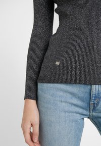 Svart ribbad stickad tröja med metalltråd, med en liten silverfärgad logotagg vid fållen, tillsammans med ljusblå denimjeans.