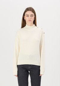 Maglione lavorato a maglia crema con una texture a coste, caratterizzato da un collo a imbuto e bottoni dorati su una spalla. Abbinato a jeans scuri.