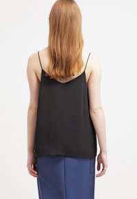 Femme aux longs cheveux roux portant un haut noir sans manches à fines bretelles et une jupe bleue, debout de dos sur un fond uni.