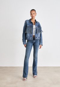 Femme debout portant une veste en jean bleue avec franges, un jean assorti, un crop top blanc et des talons ouverts argentés sur un fond uni.