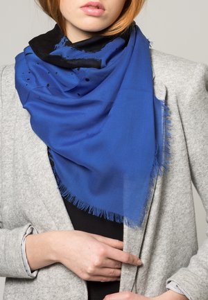 Femme portant une grande écharpe bleue et noire à franges sur un blazer gris, ajustant le blazer de la main droite.