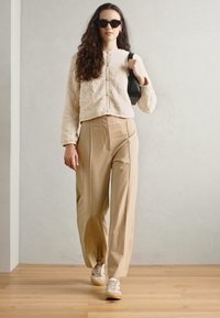 Light beige textured jacket with a zip, paired with high-waisted beige trousers, and light sneakers. Accessories include sunglasses.