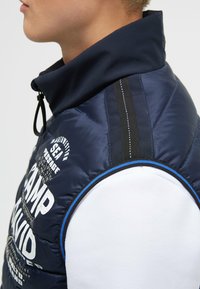 Marineblauer Steppweste mit hohem Kragen, Akzenten in Schwarz-Weiß-Streifen. Verfügt über bedruckten Text und Grafikdesign auf der linken Seite, mit blauen Trimm.