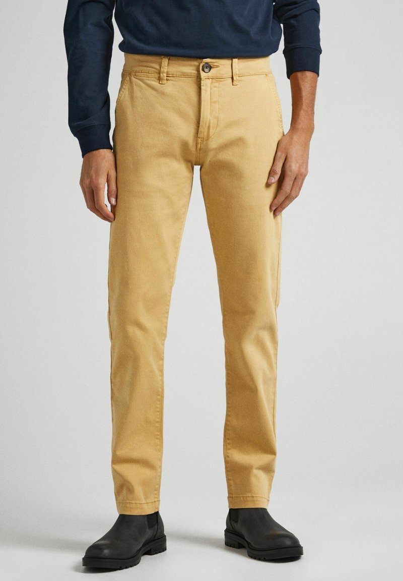 Pantalon jaune pâle en coton, avec une coupe classique, des poches avant et une seule poche arrière, associé à des bottines noires.