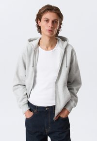 Sweat à capuche gris clair zippé avec capuche à cordon, poches avant plates, porté sur un t-shirt blanc, associé à un jean bleu foncé.