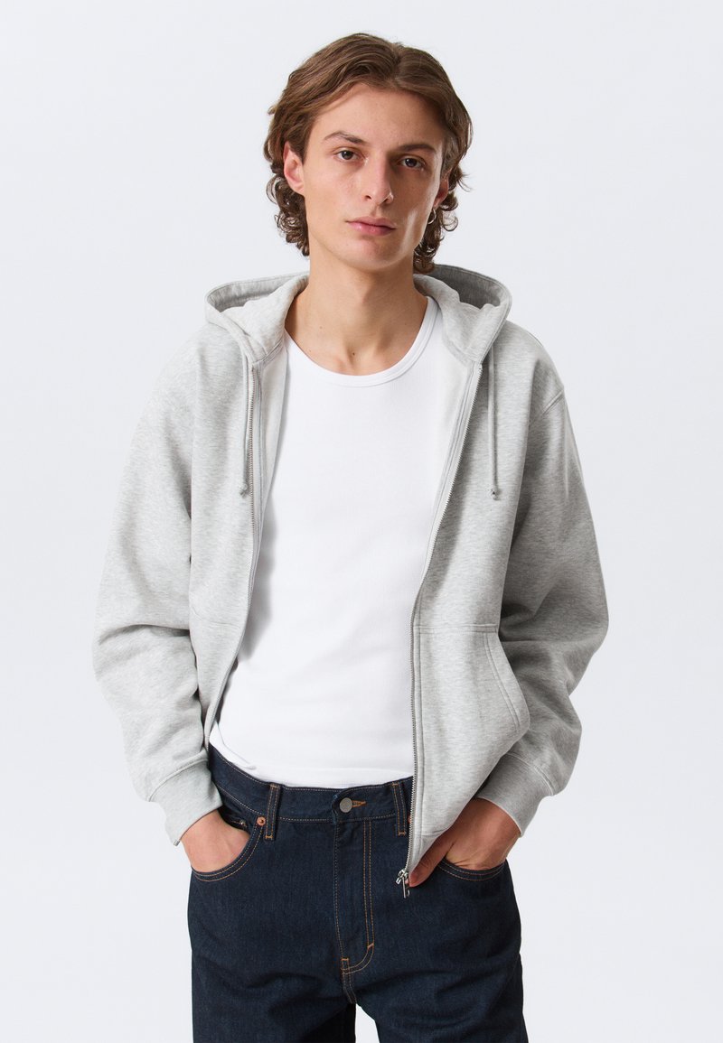 Sweat à capuche gris clair zippé avec capuche à cordon, poches avant plates, porté sur un t-shirt blanc, associé à un jean bleu foncé.