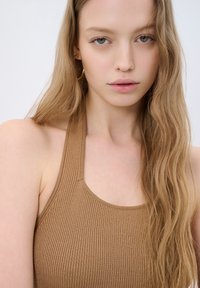 Hnědý žebrovaný halter top s širokým výstřihem. Tkanina má mírný lesk a je přiléhavá. Dlouhé vlnité vlasy rámují obličej.