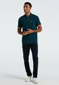 Türkises Polohemd mit Tasche und Dreiknopf-Leiste, kombiniert mit navyfarbenen Cargohosen und weißen Sneakern, stehend vor einem hellen Hintergrund.