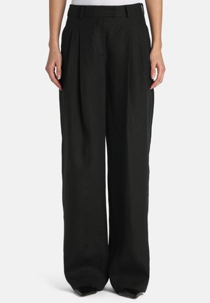 Person trägt hochtaillierte, weit geschnittene schwarze Hosen mit Falten und einem Taillenbund, kombiniert mit spitzen schwarzen Schuhen vor einem einfarbigen Hintergrund.