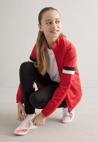 Rdeča športna jakna s črnimi in belimi črtami, kombinirana s črnimi hlačami ter belimi čevlji z oranžnimi poudarki. Poza v prikleknjenem položaju.