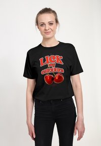 Mladá žena se světlými vlasy má na sobě černé tričko s obrázkem dvou červených třešní a textem "LICK my CHERRIES" velkými červenými písmeny.