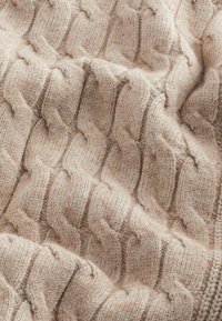 Tela de punto beige con un patrón de cable elevado, con una apariencia suave y texturizada y variaciones de tono a lo largo del tejido.