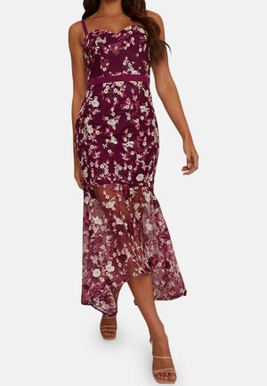 Femme portant une robe sans manches à fleurs violettes, avec un corsage ajusté et une jupe fluide et transparente, associée à des talons ouverts beiges.