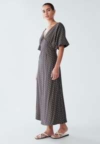 Grå maxikjole med prikker, dyb V-hals, korte flagrende ærmer og en afslappet pasform. Kombineret med minimalistiske hvide sandaler.
