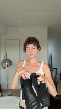 Melnās sporta kurpes, izgatavotas no gludas ādas, ir aprīkotas ar auklām, teksturētām akcentiem un mīkstu apkaklīti. Parādītas turētas rokās, tuvplāna skats.