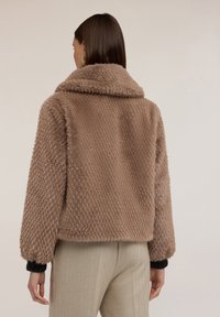 Giacca in ecopelliccia marrone con superficie testurizzata e decorata, ampio colletto e polsini aderenti. Il modello è corto e dalla forma squadrata.