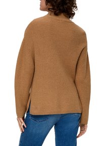 Brauner, gerippter Pullover mit hohem Kragen, fallenden Schultern und seitlichen Schlitzen. Kombiniert mit blauen Jeans, die eine lockere Passform zeigen. Die Textur ist gestrickt.